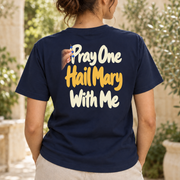Woman wearing the navy Pray One Hail Mary With Me T-shirt in a garden, showing the updated tall cream-and-gold back print and small hand holding blue rosary beads.