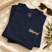 Folded navy Pray One Hail Mary With Me T-Shirt showing the ivory and gold Let's pray! print beside sunglasses and olive branches.